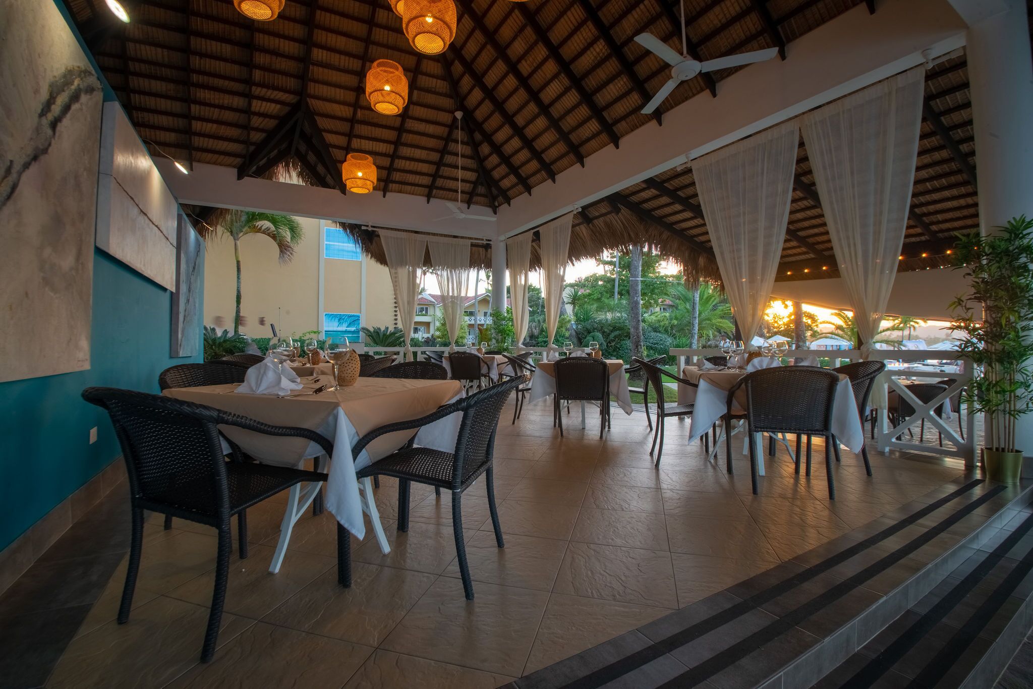 An outdoor dining area with wicker chairs and tables, a thatched roof, and curtains.