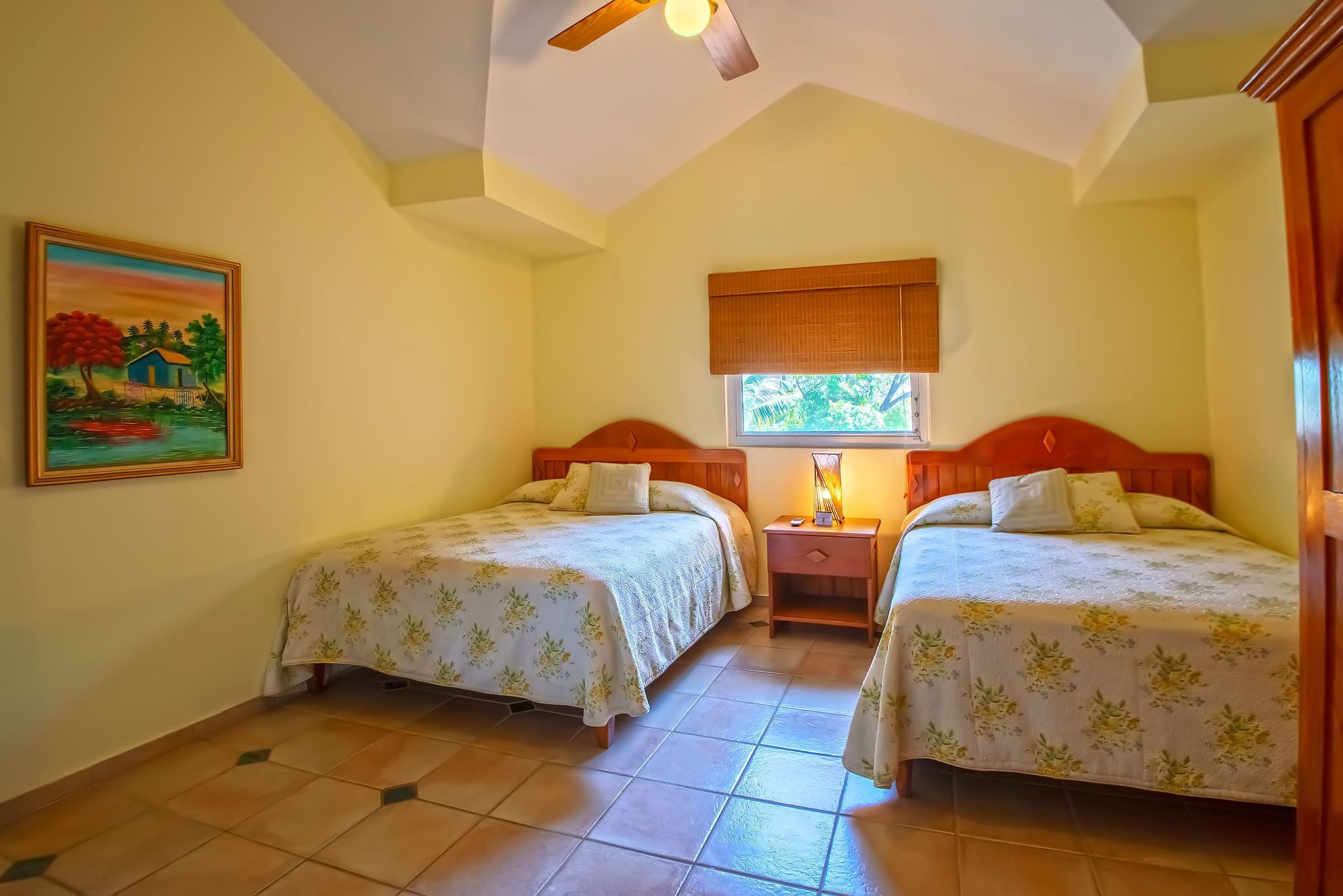 Une chambre avec deux lits, un ventilateur de plafond, une fenêtre avec un store et un tableau au mur.
