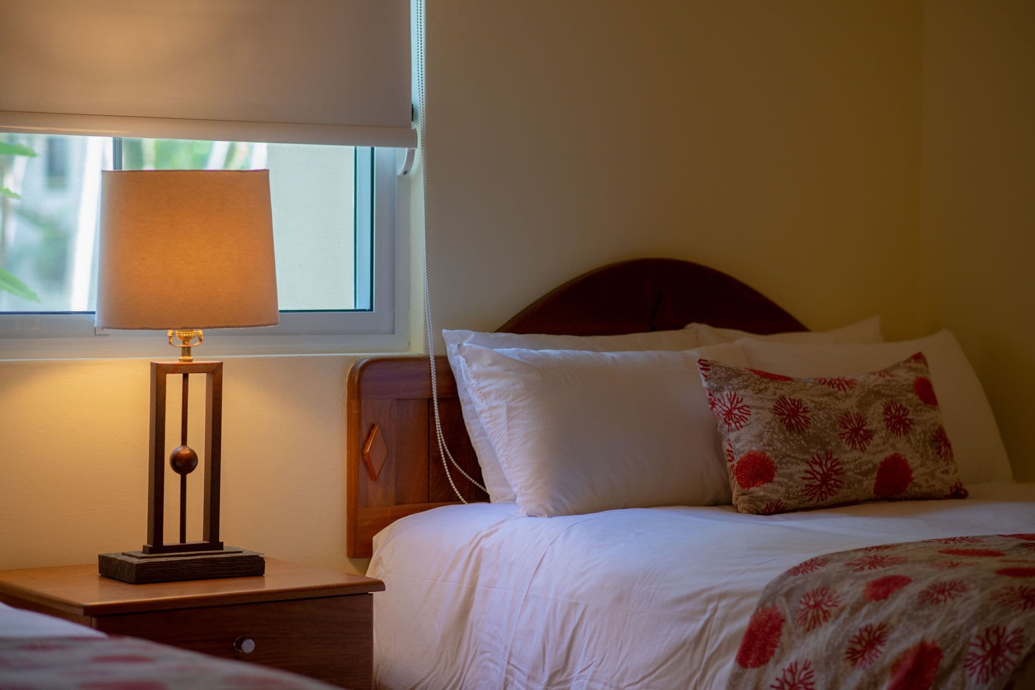 Una cama bien hecha con ropa de cama blanca y una manta decorativa de estampado floral, una mesita de noche con una lámpara, y una ventana con una persiana bajada a medias.