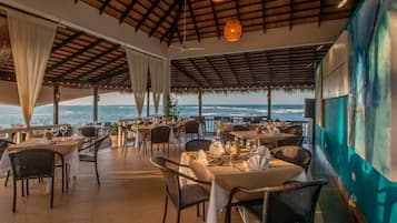 An outdoor dining area with tables and chairs, a ceiling with hanging lights, and a view of the ocean.