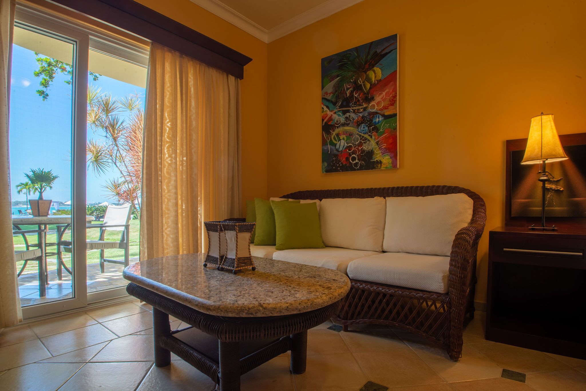 A room with a wicker sofa, a round table, a painting on the wall, and a view through a glass door.