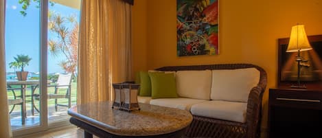 A room with a wicker sofa, a round table, a painting on the wall, and a view through a glass door.