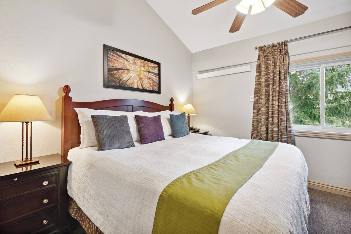 A neatly made bed with a wooden headboard, two bedside lamps, a framed artwork above the bed, and a window with curtains.