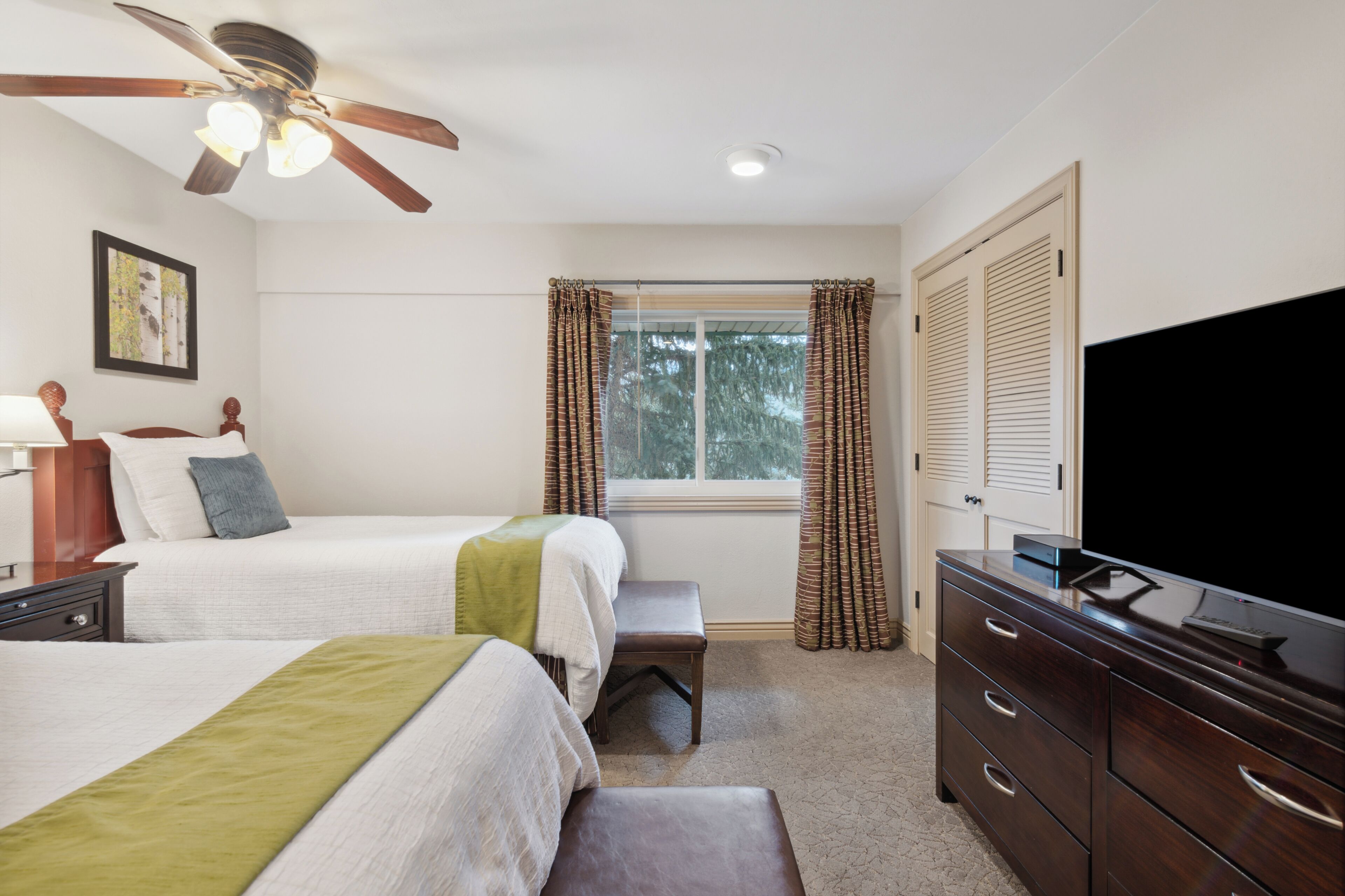 A hotel room with two beds, a ceiling fan, a television, a dresser, and a window with curtains.