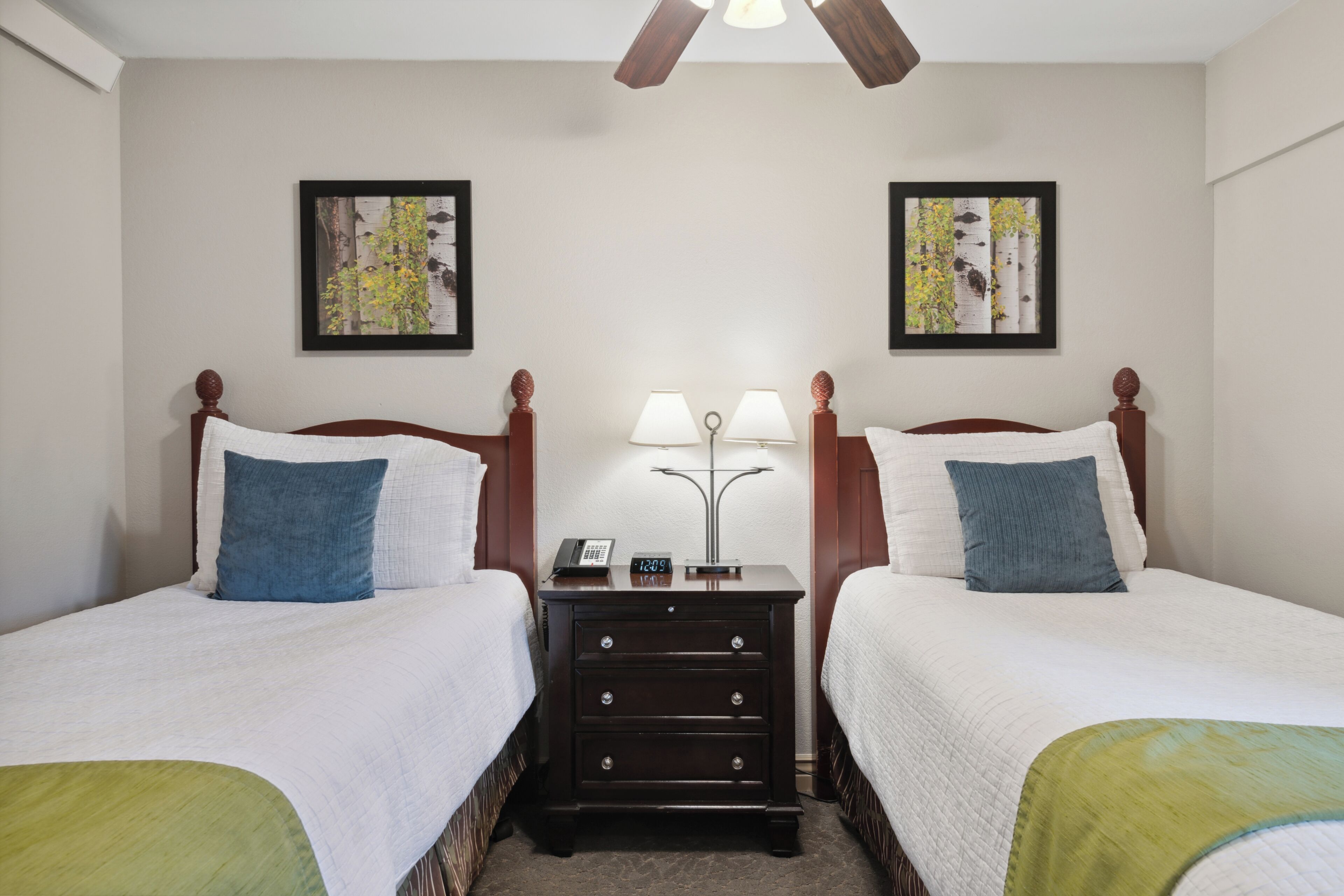 Habitación de hotel con dos camas, cada una con cabecera de madera, ropa de cama blanca y un plaid verde al pie. Hay una mesita de noche entre las camas con una lámpara y un teléfono.