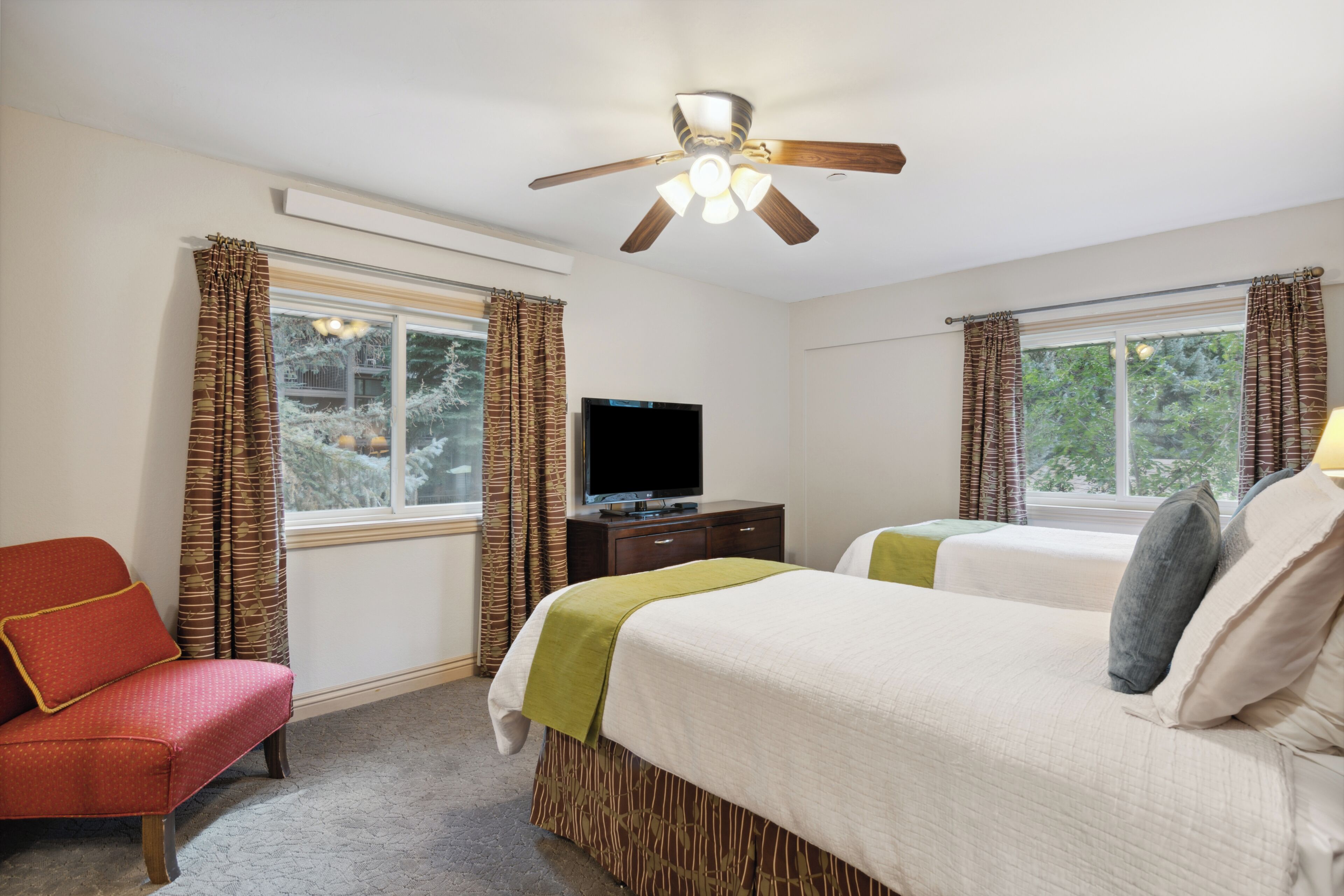 A hotel room with two beds, a television, a ceiling fan, and a window with curtains.