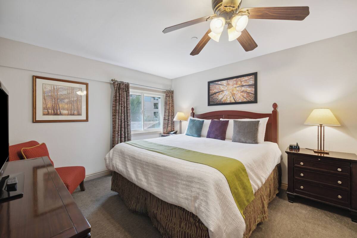 A hotel room with a large bed, a wooden headboard, a ceiling fan, a nightstand with a lamp, a framed picture on the wall, and a window with curtains.