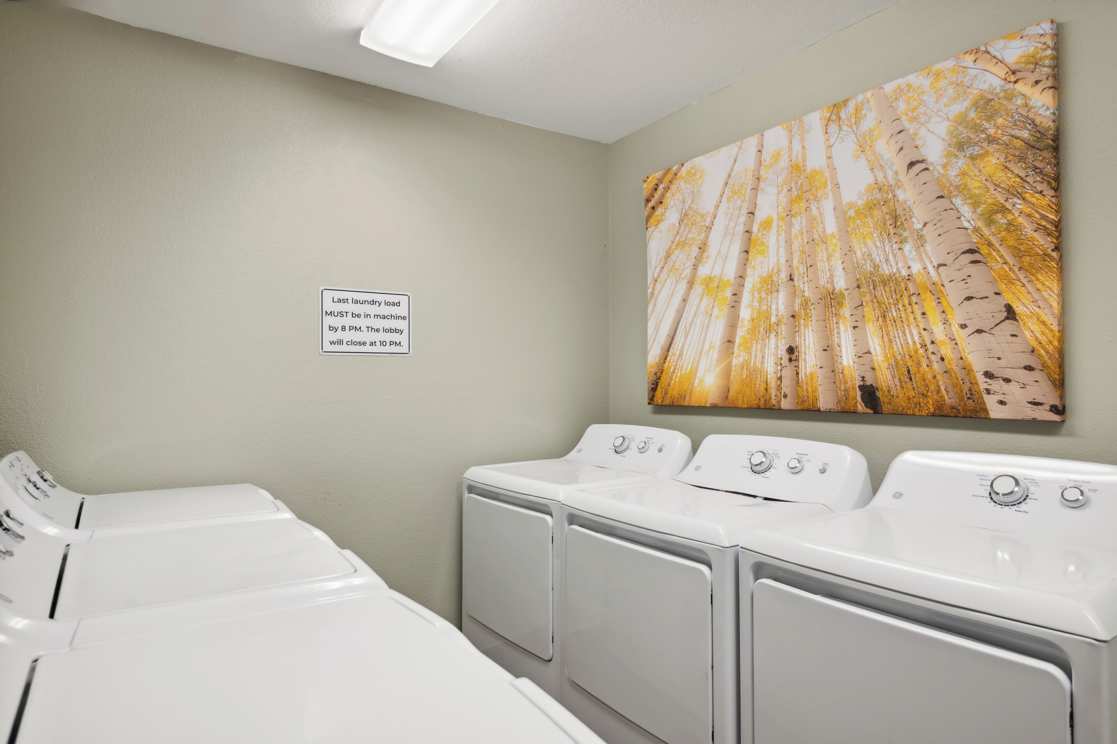 A laundry room with four white washers and dryers, a sign indicating the last load must be in by 8 PM, and a large painting of birch trees on the wall.