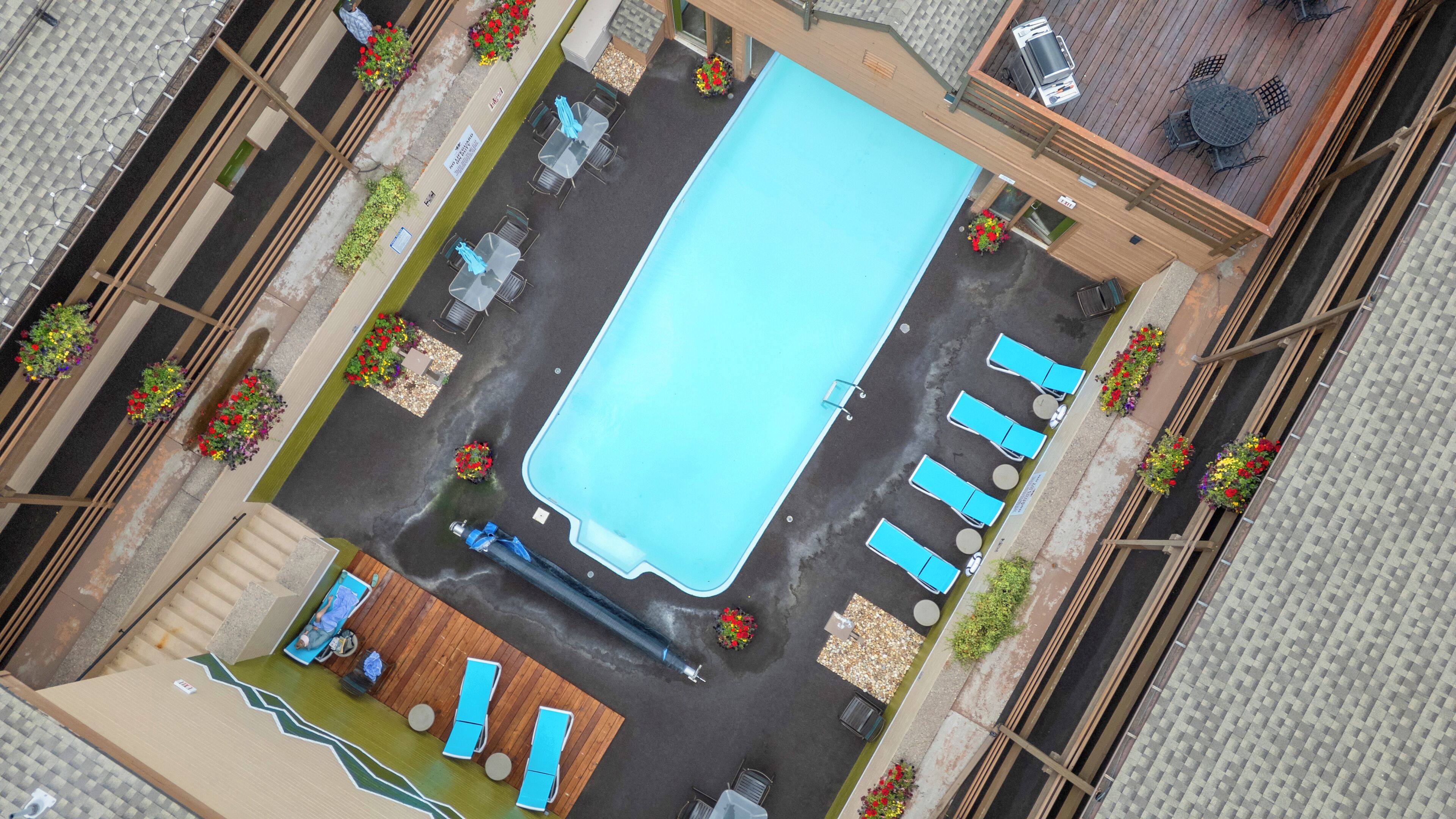 An aerial view of a pool area with lounge chairs, a small deck, and surrounding flower beds.