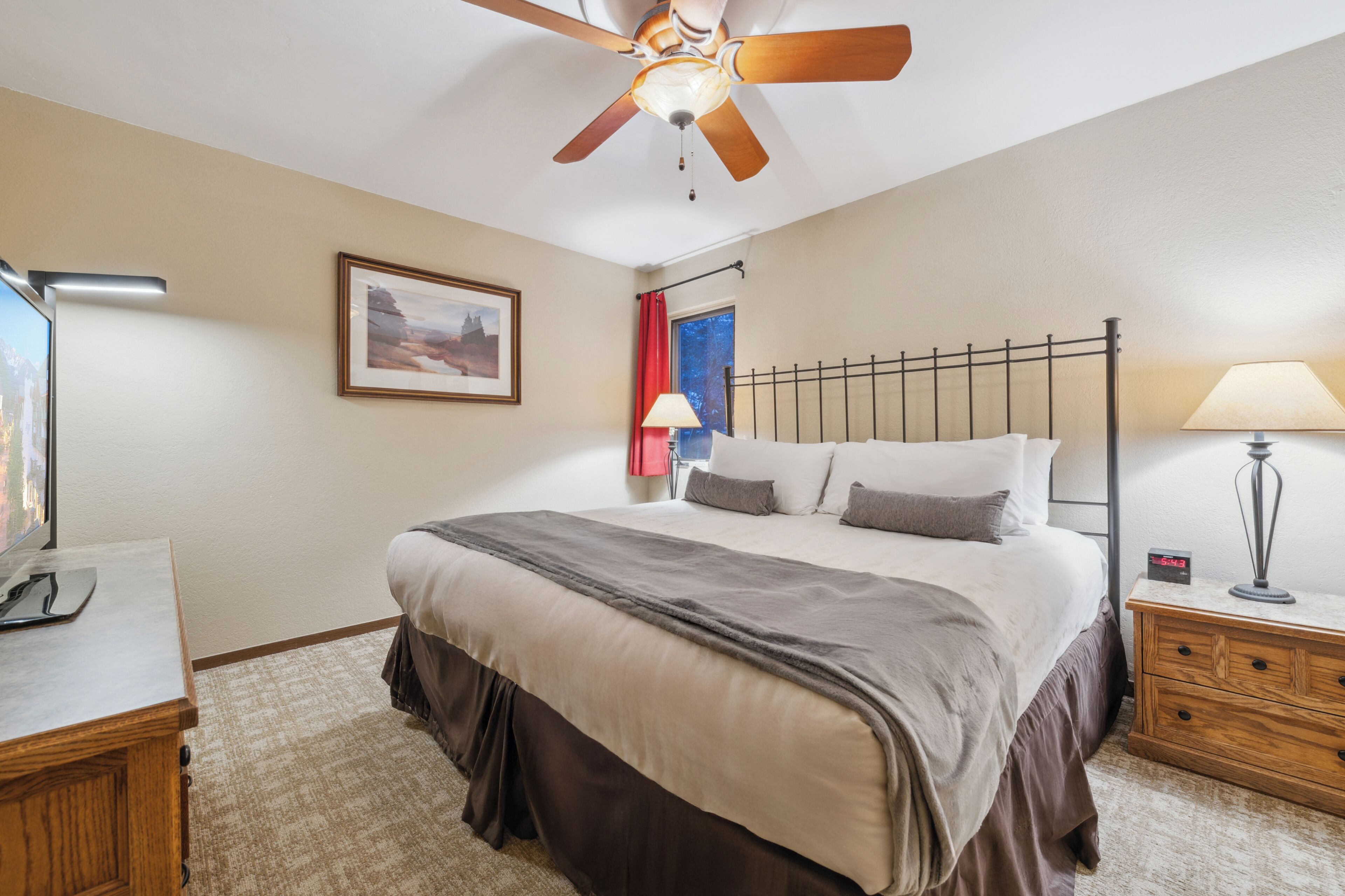 A bedroom with a bed, a ceiling fan, a nightstand, a lamp, a framed picture, and a window with a red curtain.