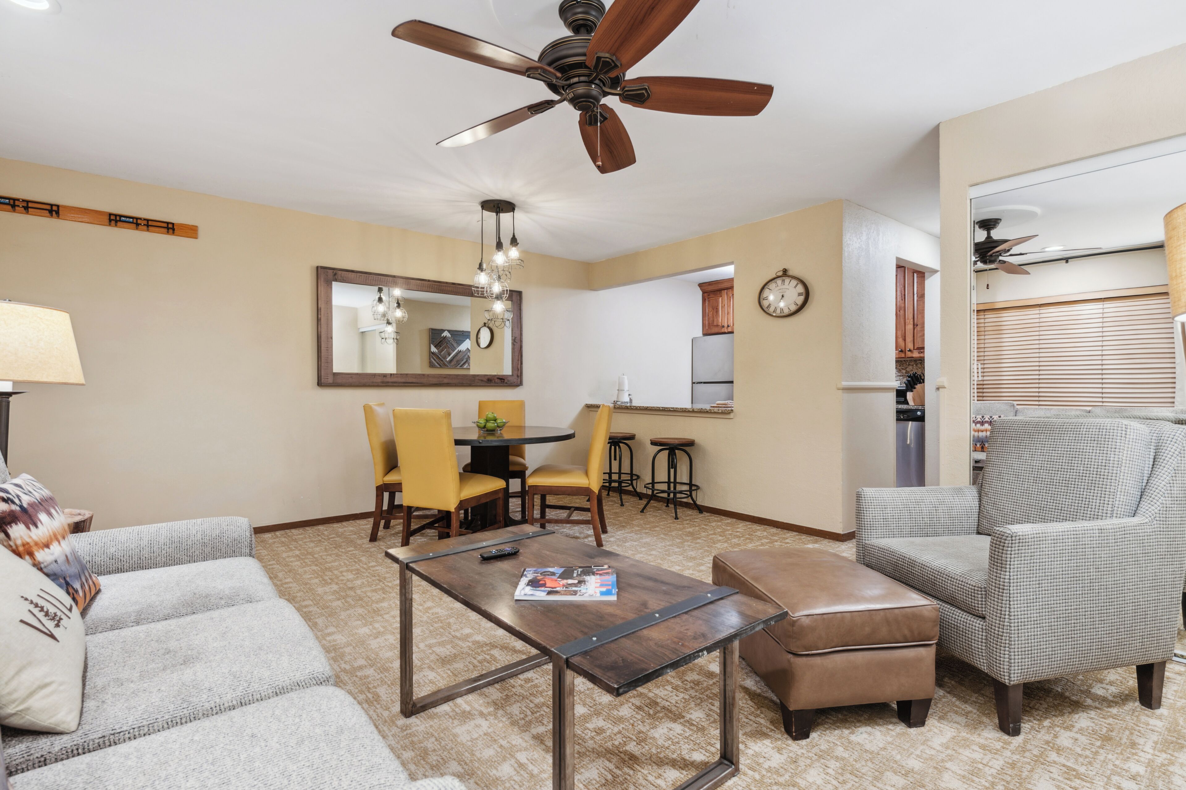 Sala de estar con sofá, reposapiés, mesa de centro y ventilador de techo. Hay un comedor con sillas y una mesa, y una cocina con refrigerador y un reloj.