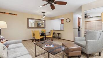 Sala de estar con sofá, reposapiés, mesa de centro y ventilador de techo. Hay un comedor con sillas y una mesa, y una cocina con refrigerador y un reloj.