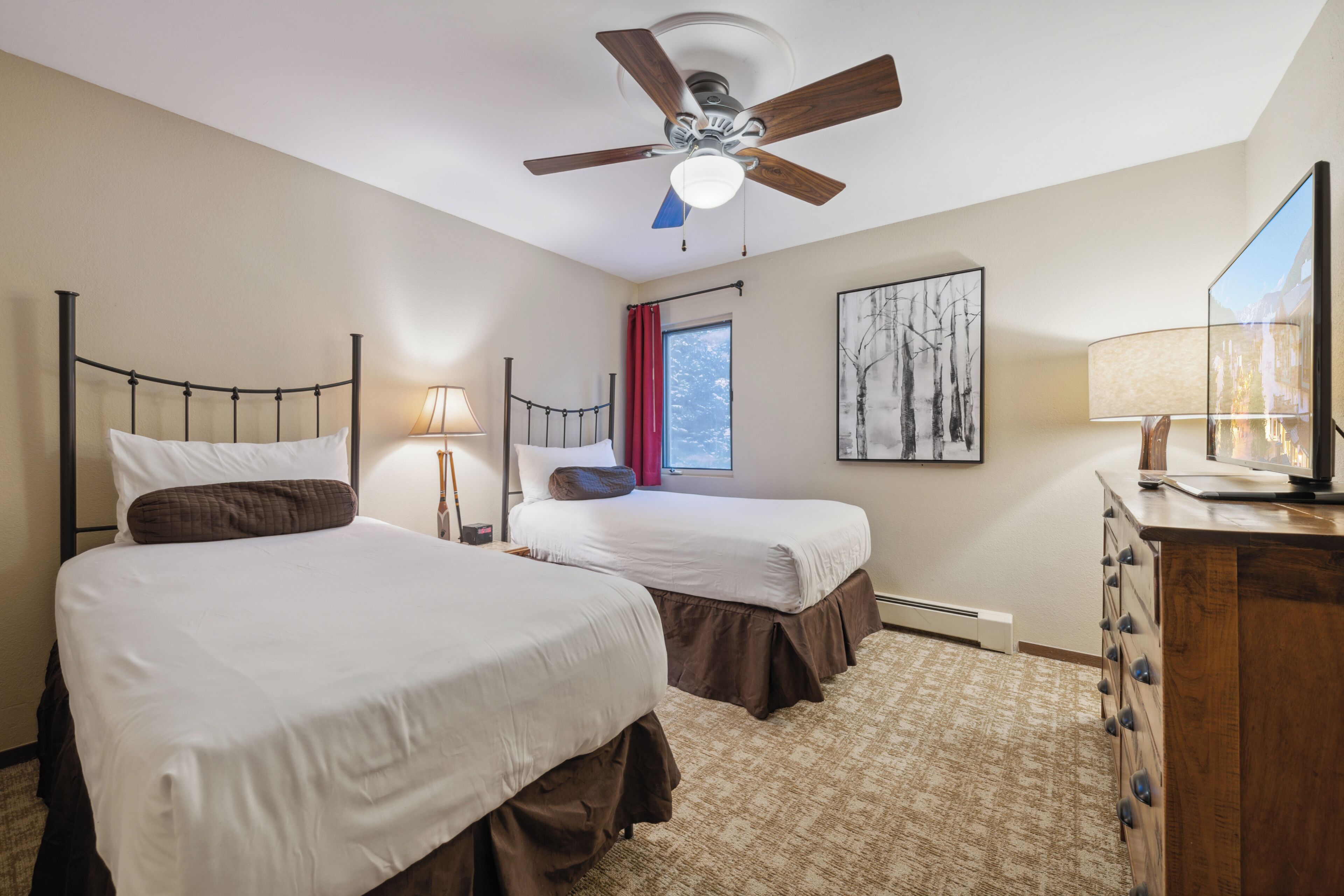 A hotel room with two beds, a ceiling fan, a window with red curtains, a lamp, and a dresser.
