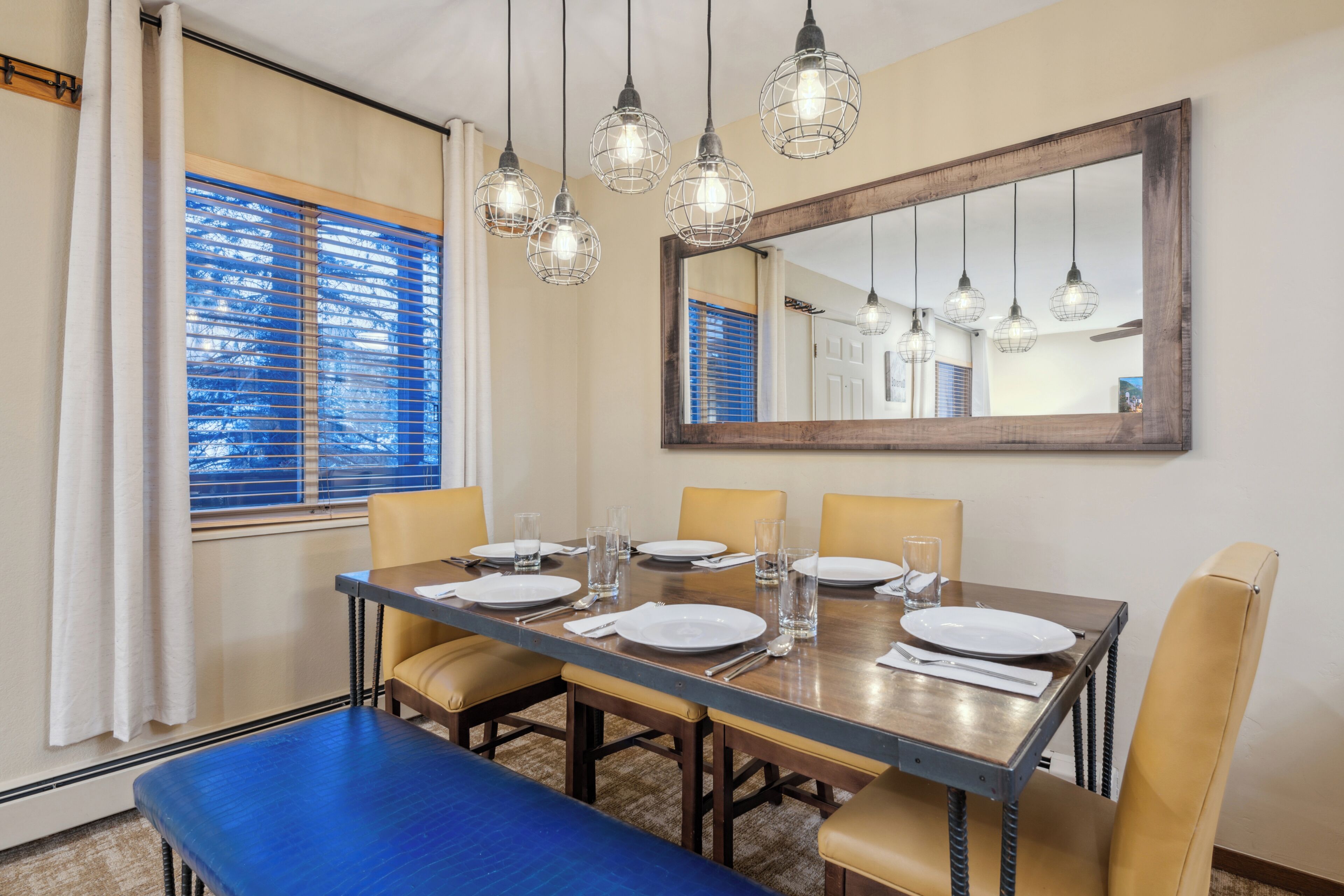 A dining area with a long table set for four, a large mirror above the table, and hanging pendant lights.