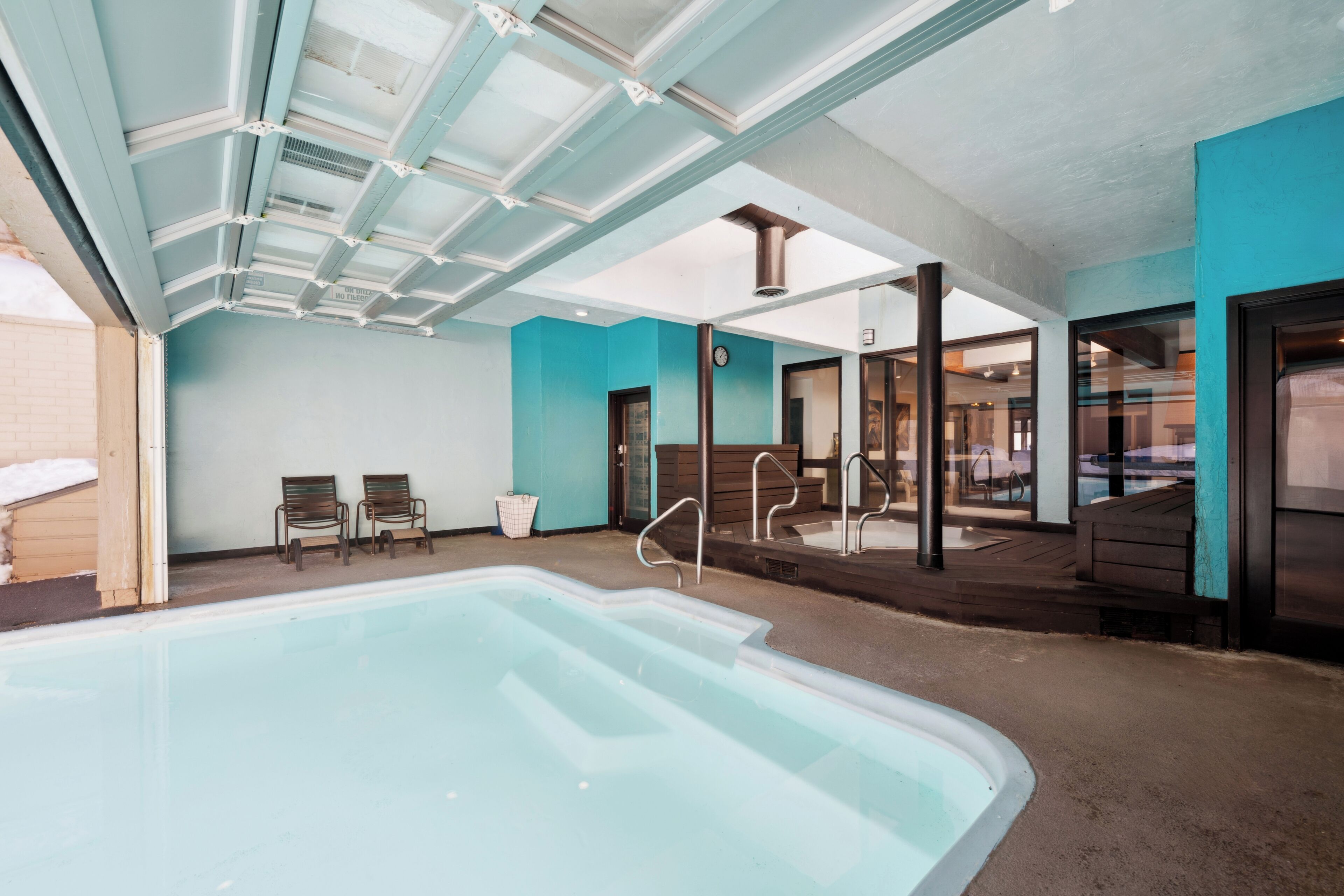 Une piscine intérieure avec des parois en verre transparent, une terrasse en bois et deux chaises longues.