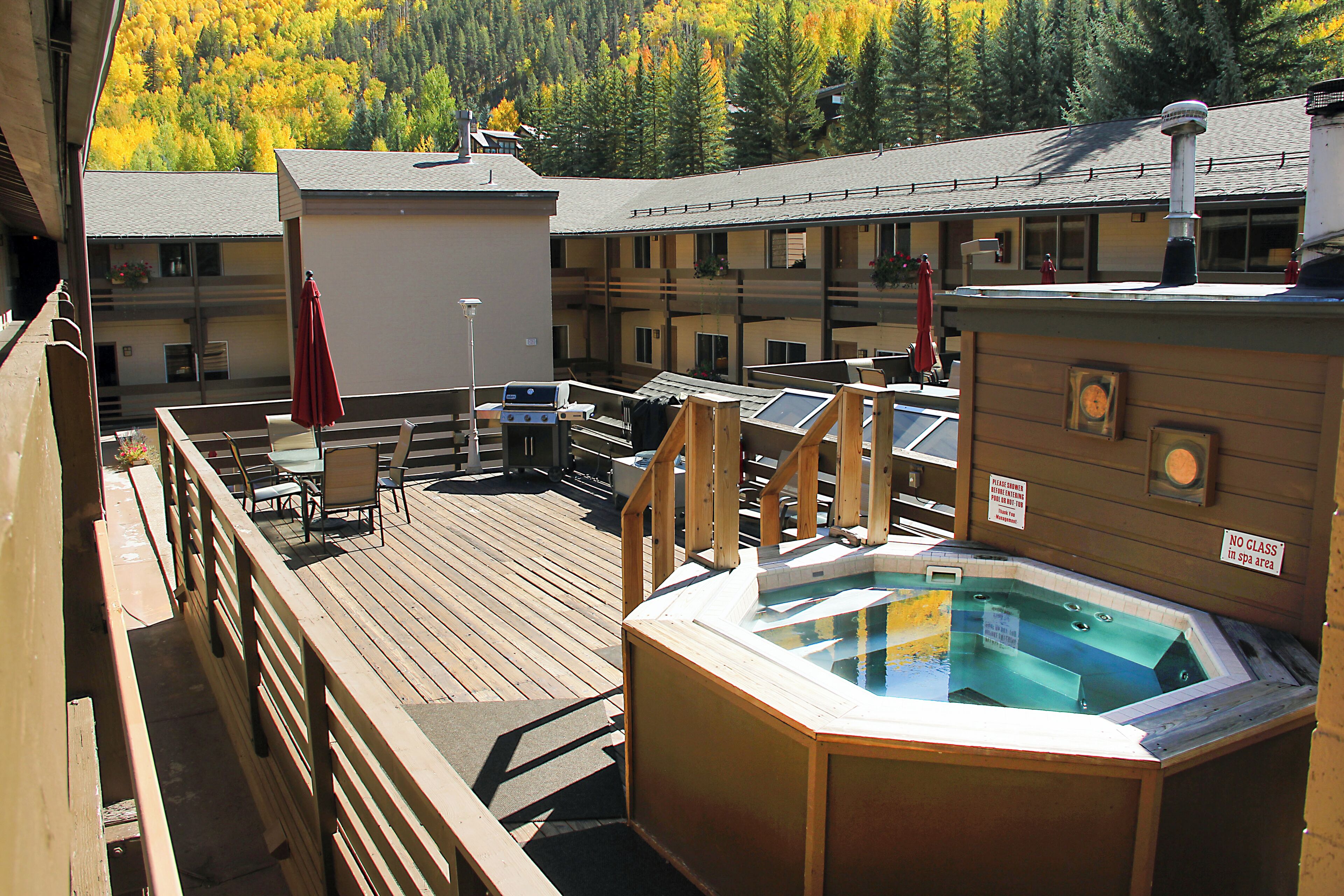 A wooden deck with a hot tub, outdoor seating, and a view of autumn trees.