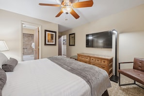 A hotel room with a large bed, a wooden dresser, a leather chair, a ceiling fan, and a flat-screen TV mounted on the wall.