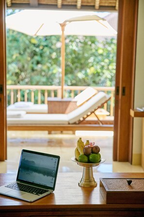A laptop on a wooden table with a fruit bowl in the foreground and a balcony with lounge chairs in the background.