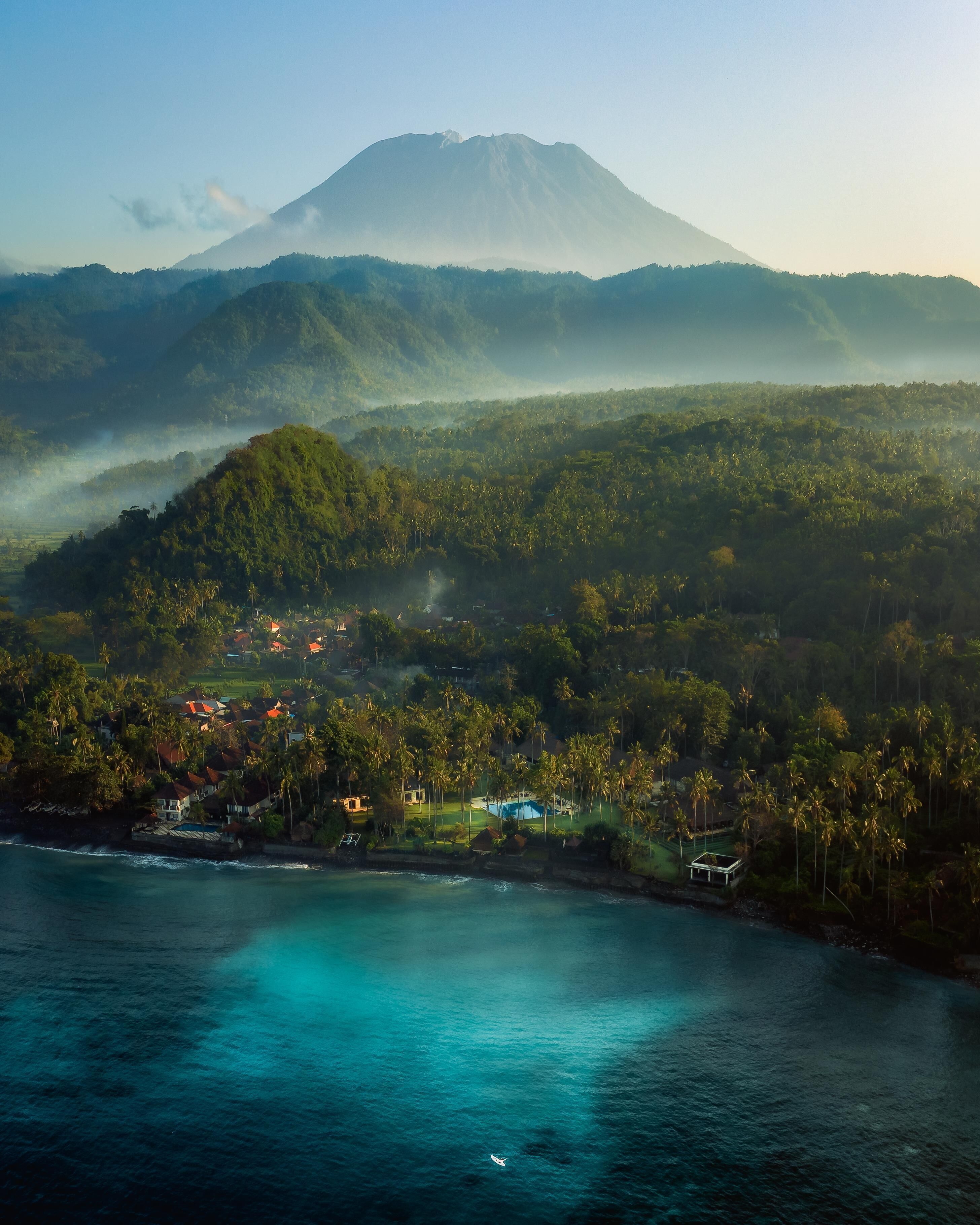 An aerial view of a coastal area with a mountain in the background, a beach with clear turquoise waters, and a cluster of houses surrounded by lush greenery.