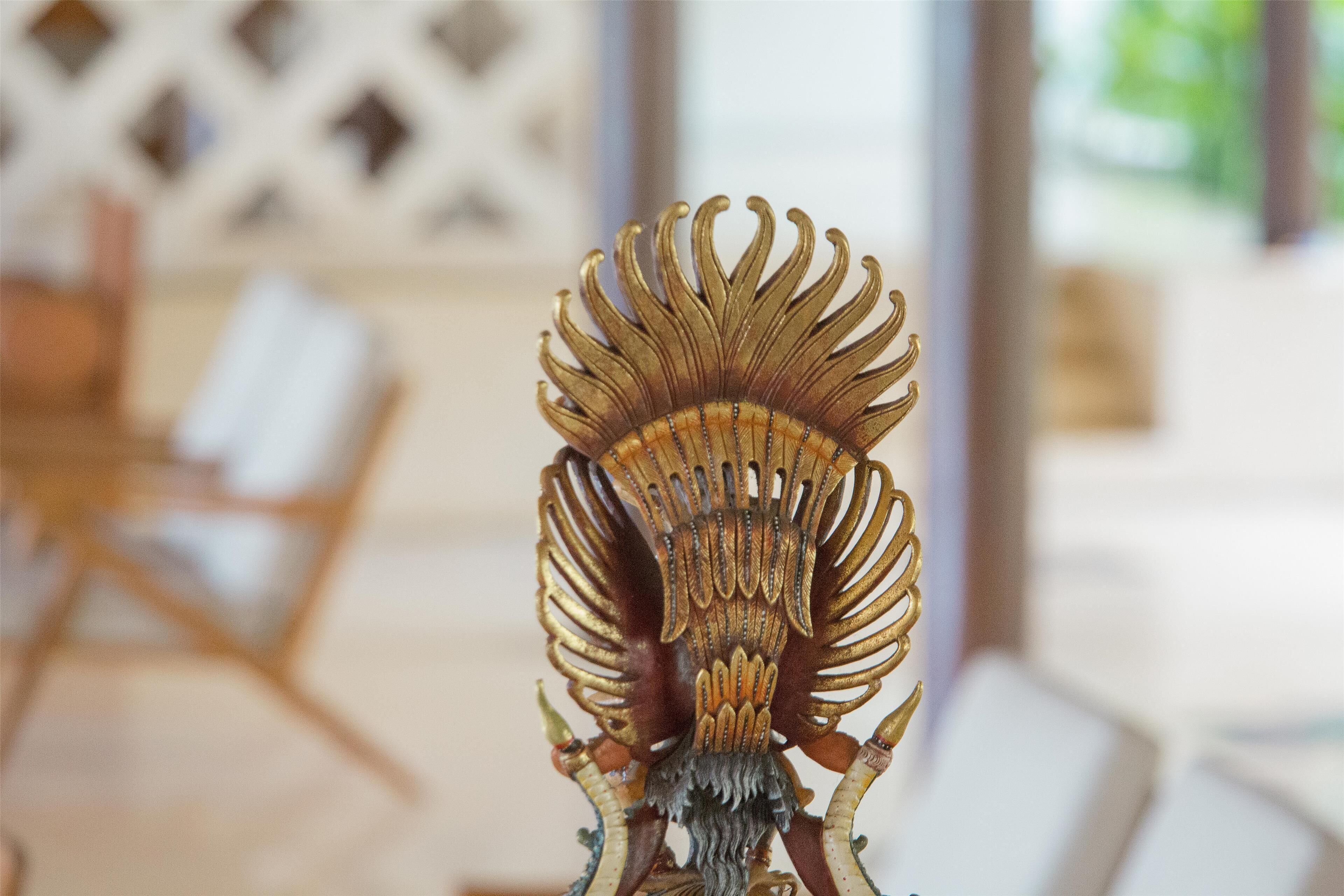 Un objeto decorativo dorado con detalles intrincados, posiblemente un mueble o una instalación artística, en una habitación con una silla blanca y un muro ajedreado al fondo.