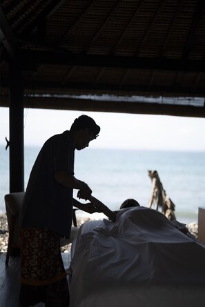 A person attending to another individual in a covered outdoor area with a view of the sea.