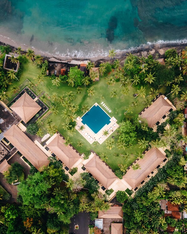 An aerial view of a resort with a central swimming pool, surrounded by lush greenery and palm trees, with a beach and ocean in the background.