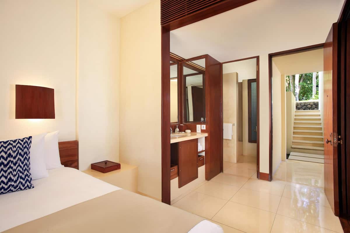 A hotel room with a bed, bedside table, and a mirror. There is a bathroom visible through an open door.