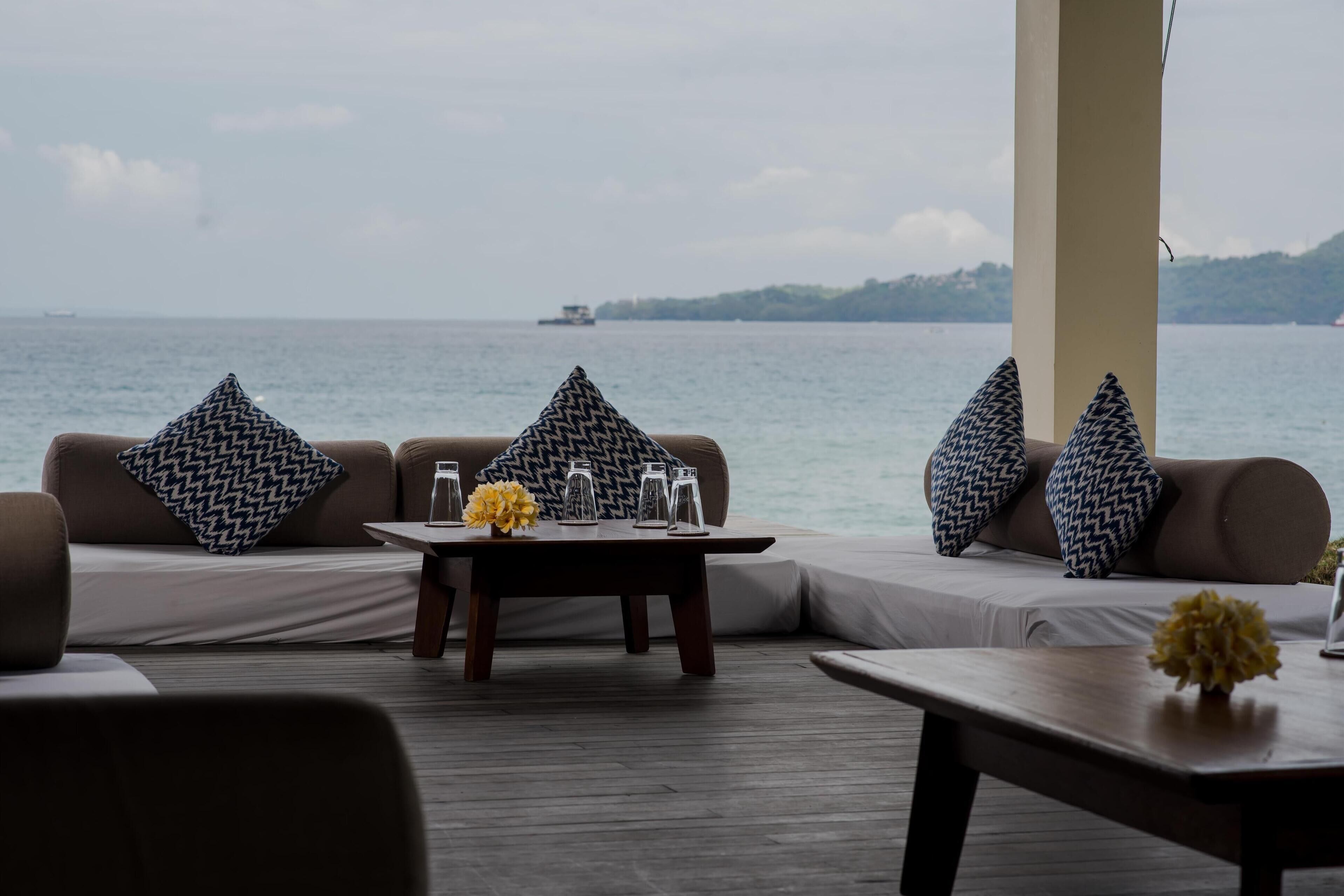 Un área de estar con vista al mar, que incluye un conjunto de sofás con cojines estampados, una mesa de madera con vasos y un florero, y piso de madera.
