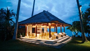 A thatched-roof outdoor lounge area with seating, surrounded by palm trees and overlooking a body of water. - Alila Manggis, Bali (Manggis)