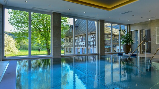 Une grande piscine intérieure offrant une vue dégagée sur une maison traditionnelle à colombages et sur la verdure environnante.