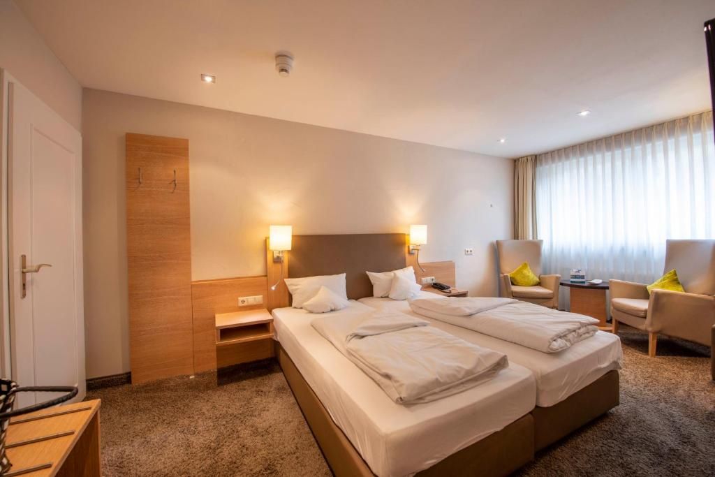 A hotel room with two beds, a wooden headboard, bedside tables, a chair, and a window with curtains.