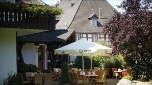 Un espace extérieur aménagé avec des tables et des chaises sous un grand parasol, un bâtiment avec un toit en pente en arrière-plan et un jardin bien entretenu.