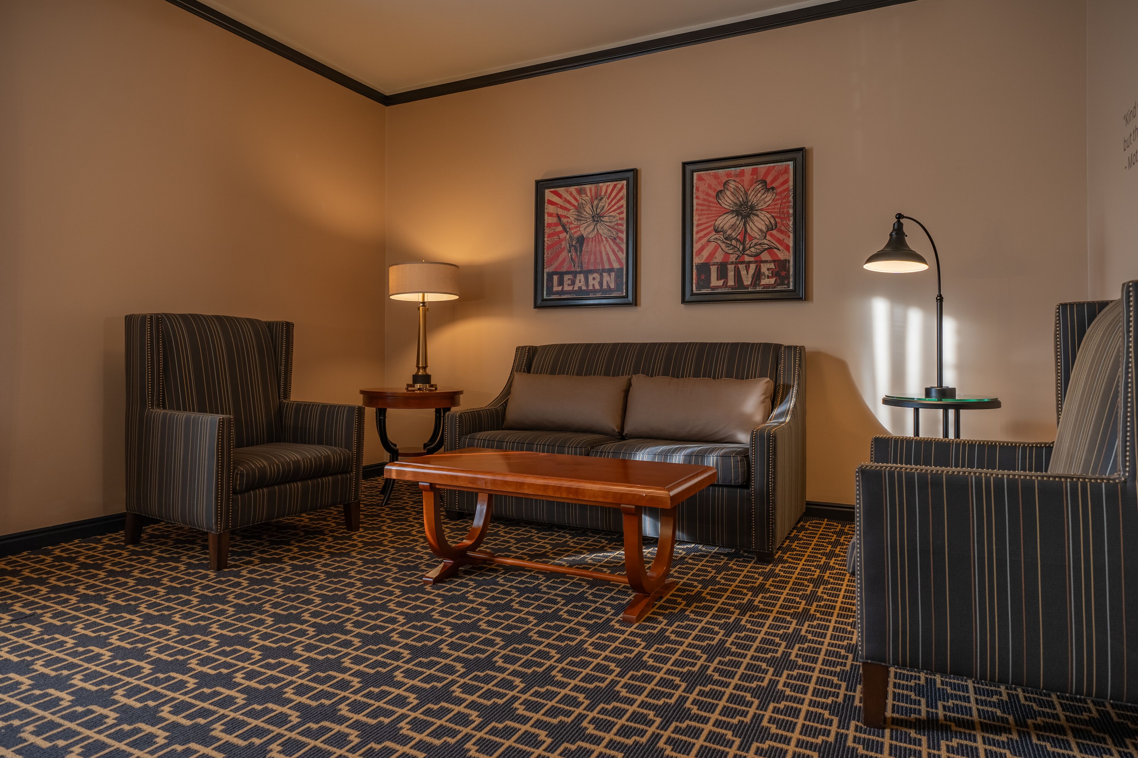Quarto de hotel com uma poltrona listrada, um sofá, uma mesa de centro de madeira e duas fotos emolduradas na parede.