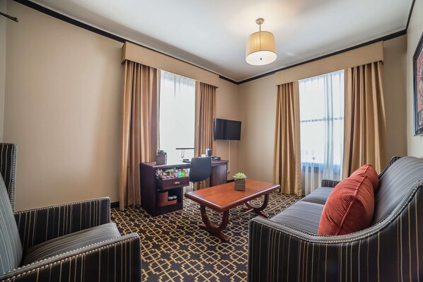 Habitación de hotel con sofá, sillones, una mesa de centro de madera, un escritorio con televisor de pantalla plana y una alfombra estampada.
