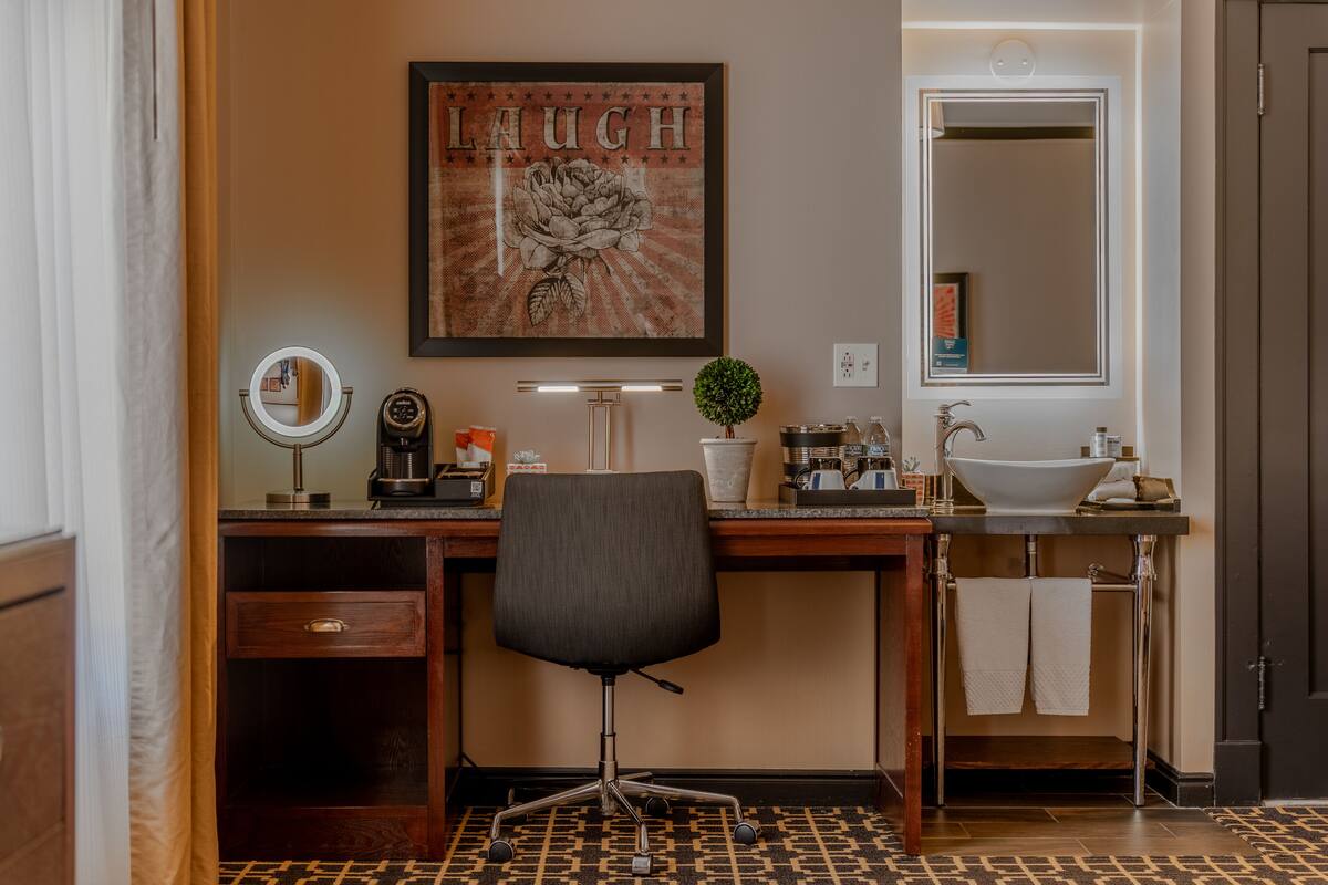 Une salle de bain avec un bureau en bois, un miroir rond, un lavabo et une œuvre d’art encadrée au-dessus du miroir.