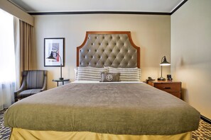 Une chambre d’hôtel avec une tête de lit capitonnée, un fauteuil à rayures, une table de chevet avec une lampe et un tableau encadré au mur.