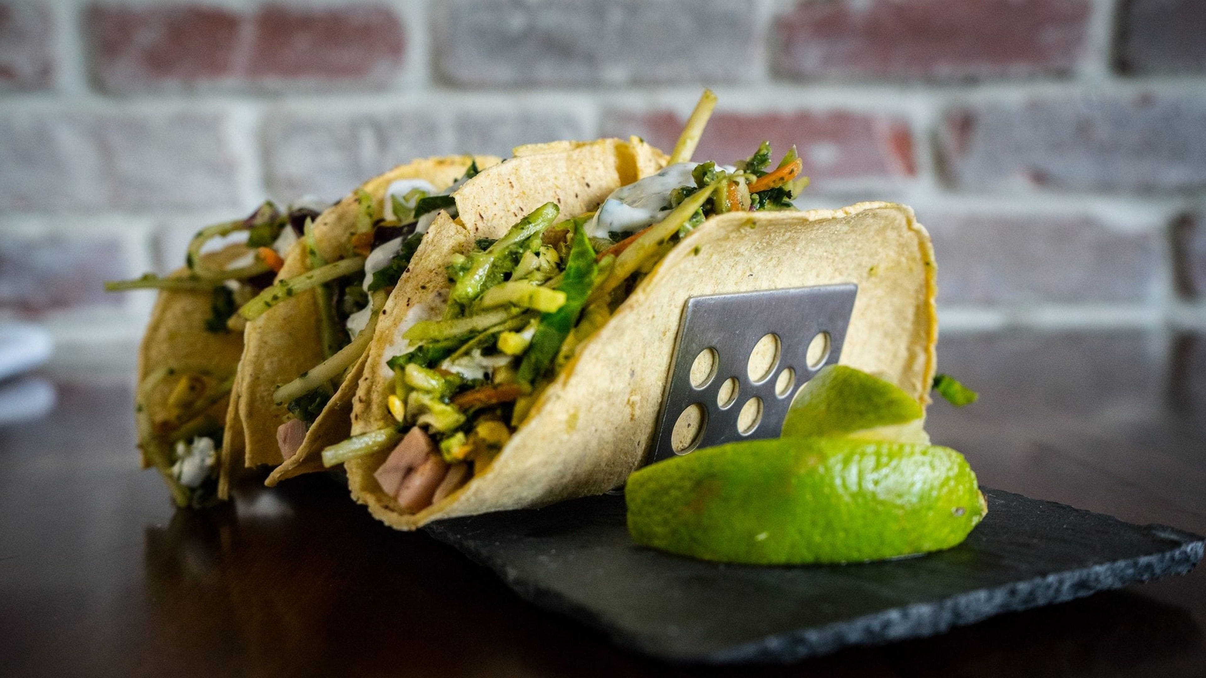 Tres tacos con una cuña de lima sobre un plato de pizarra, colocados sobre una mesa de madera con un fondo de pared de ladrillo.