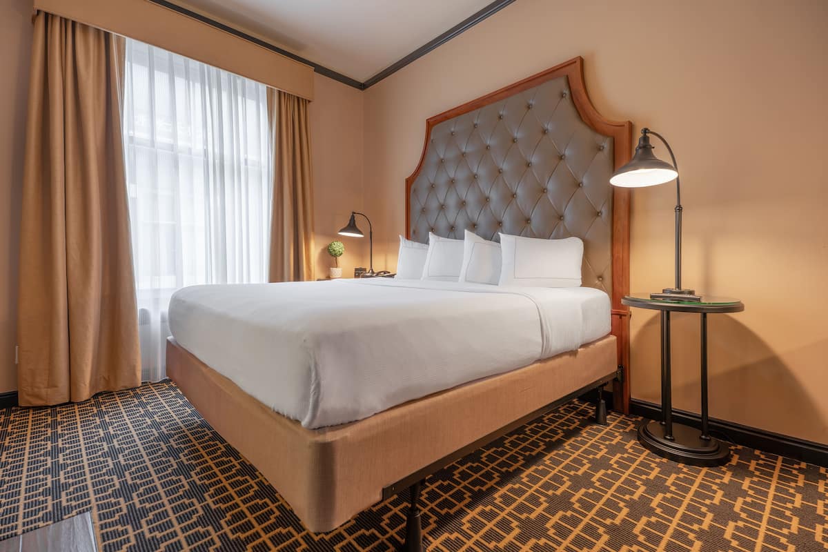 Une chambre d’hôtel avec un grand lit, une petite table ronde, une lampe et une fenêtre avec des rideaux.