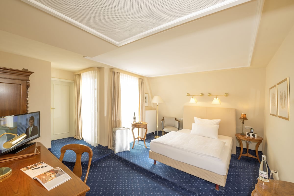 A hotel room with a bed, a desk with a computer, a chair, a small table, and a window with curtains.