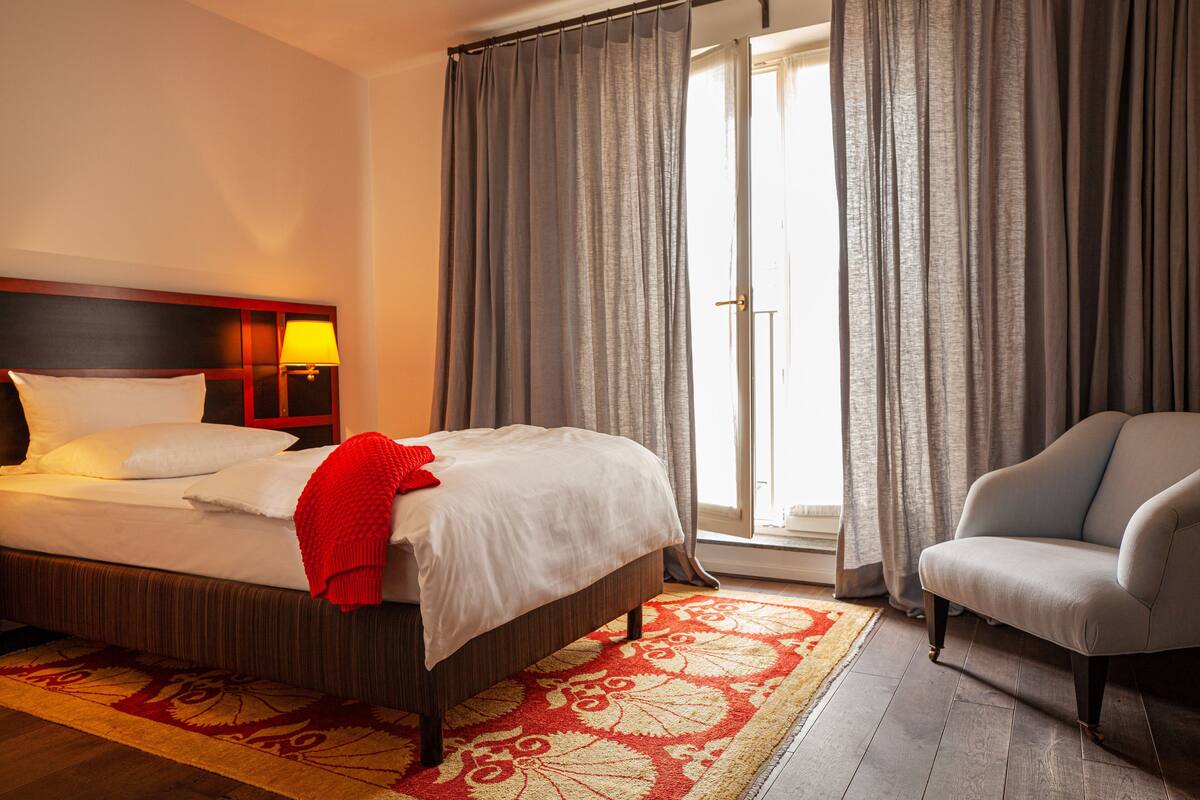 A hotel room with a bed, a chair, a nightstand, and a window with curtains.