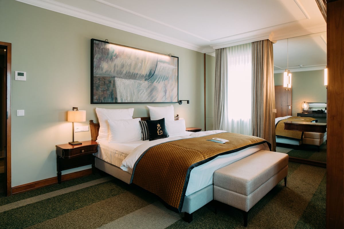 A hotel room with a large bed, a bedside table with a lamp, a bench at the foot of the bed, a large abstract painting on the wall, and a mirror on the opposite wall.