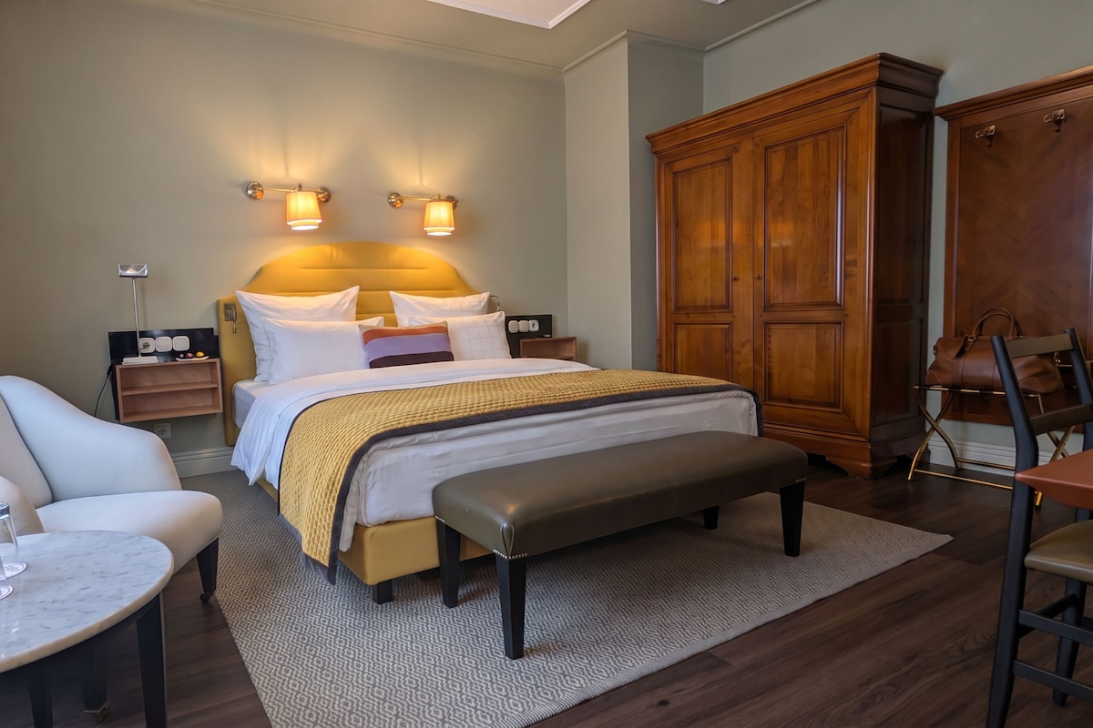 A hotel room with a bed, a desk, a chair, a wardrobe, and a bedside table with a bottle and glasses.