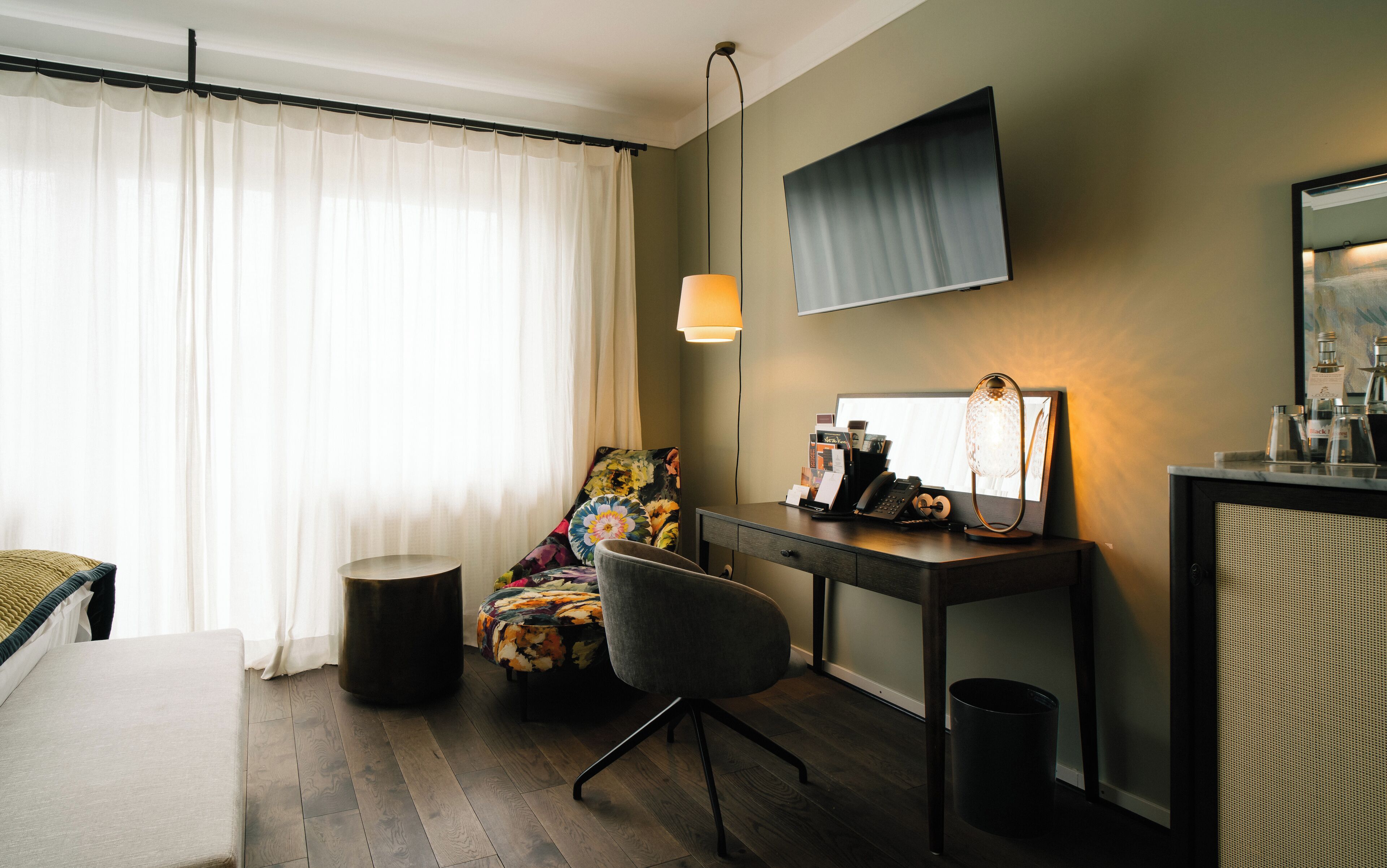 A hotel room with a bed, a desk with a lamp, a chair, a small round table, a waste bin, and a large window with curtains.