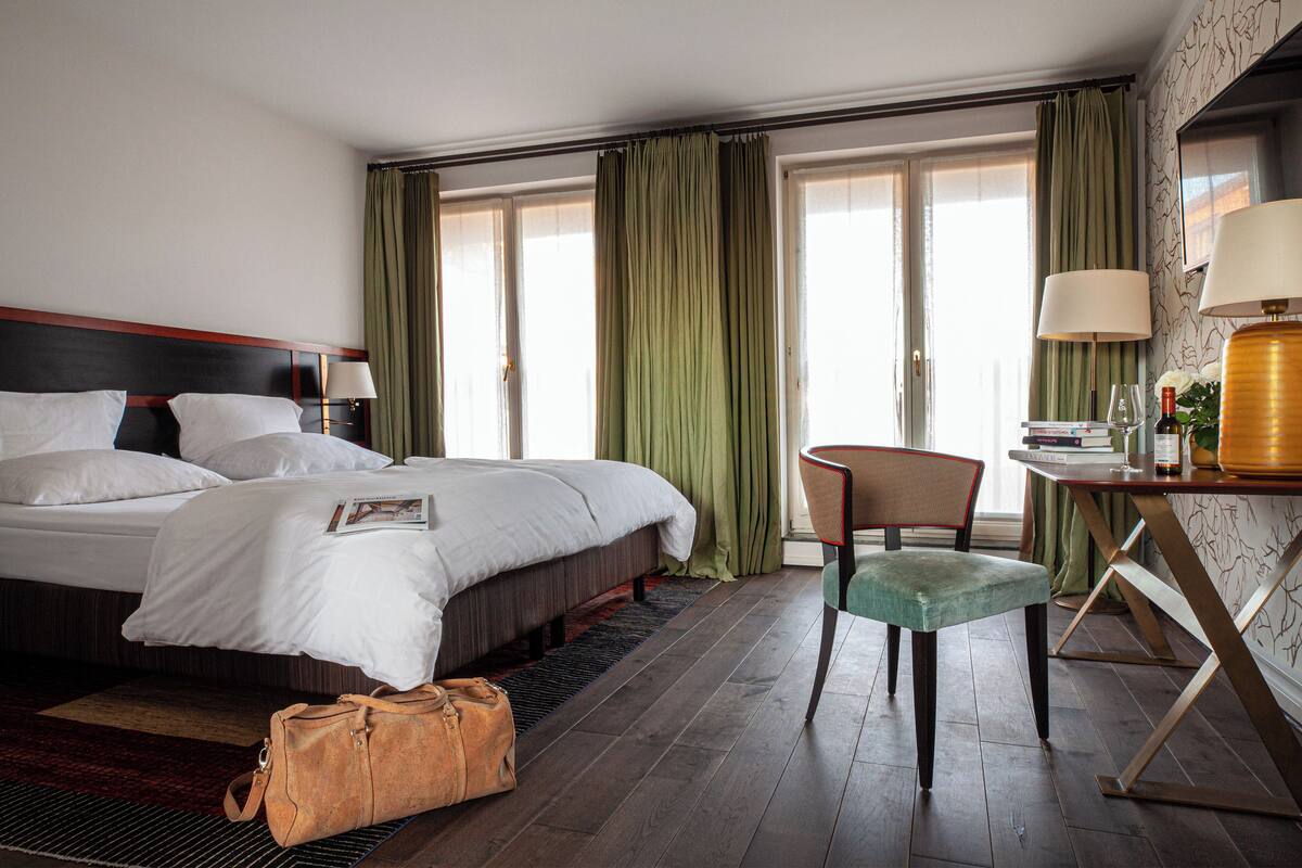 A hotel room with a large bed, a chair, a desk with a lamp, and a window with curtains.