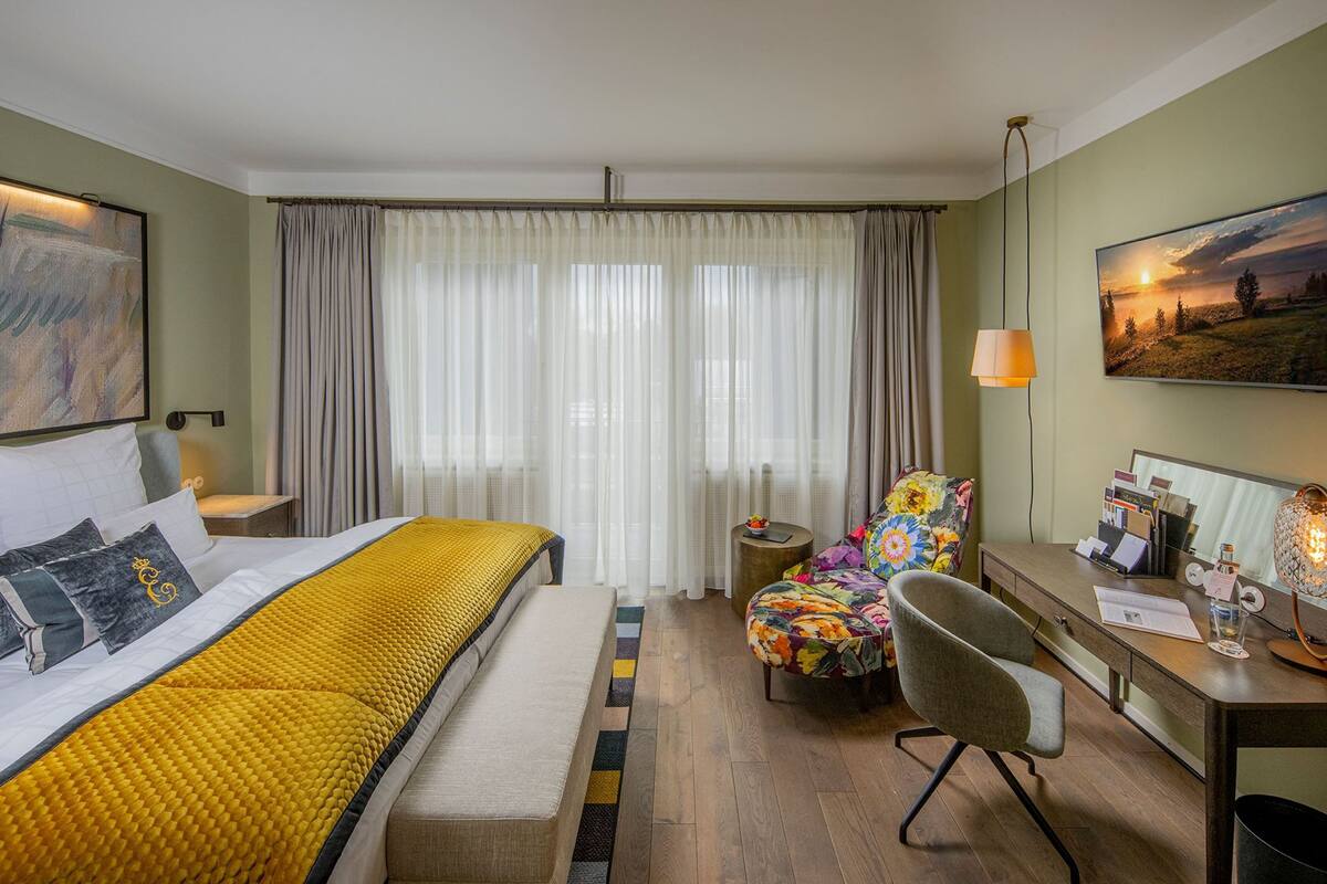 A hotel room with a bed, a desk, a chair, a small table, and a painting on the wall.