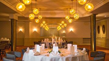 A banquet hall with round tables set for dining, surrounded by chairs, and illuminated by multiple hanging spherical lights.
