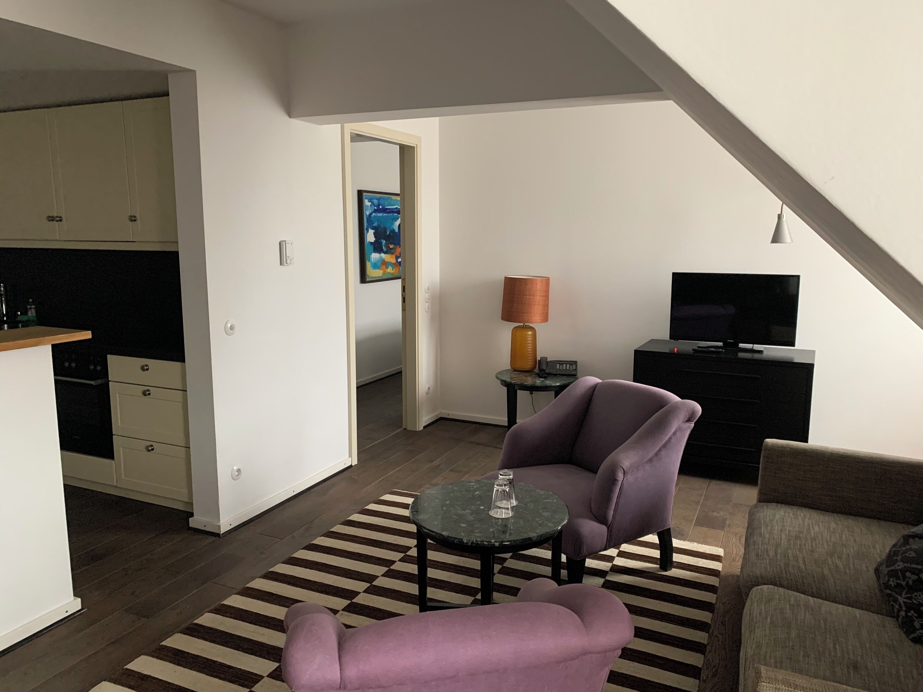 A modern living space with a kitchen, a dining area with a checkered rug, a purple armchair, a small round table, a lamp, and a television.