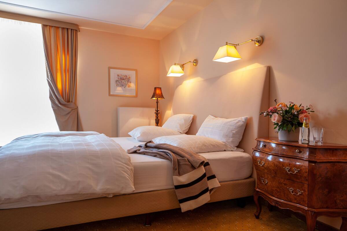 A hotel room with a bed, a dresser, a lamp, and a vase with flowers.