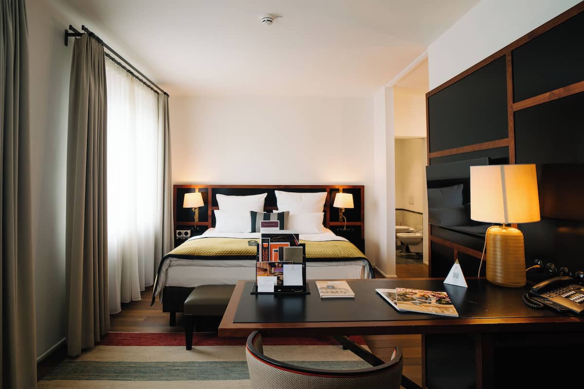 A hotel room with a bed, bedside lamps, a desk with a lamp, a chair, and a large mirror.