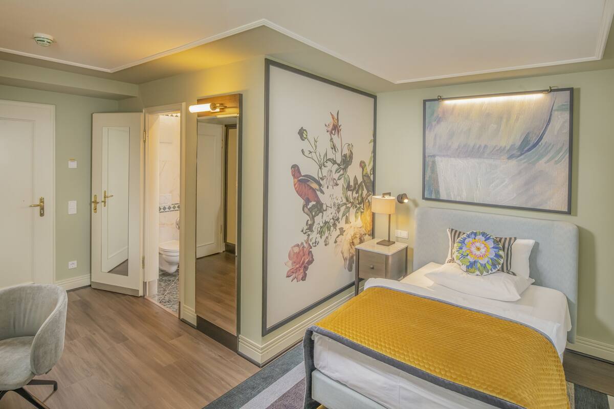 A hotel room with a bed, a chair, a nightstand with a lamp, a painting on the wall, and a door leading to the bathroom.